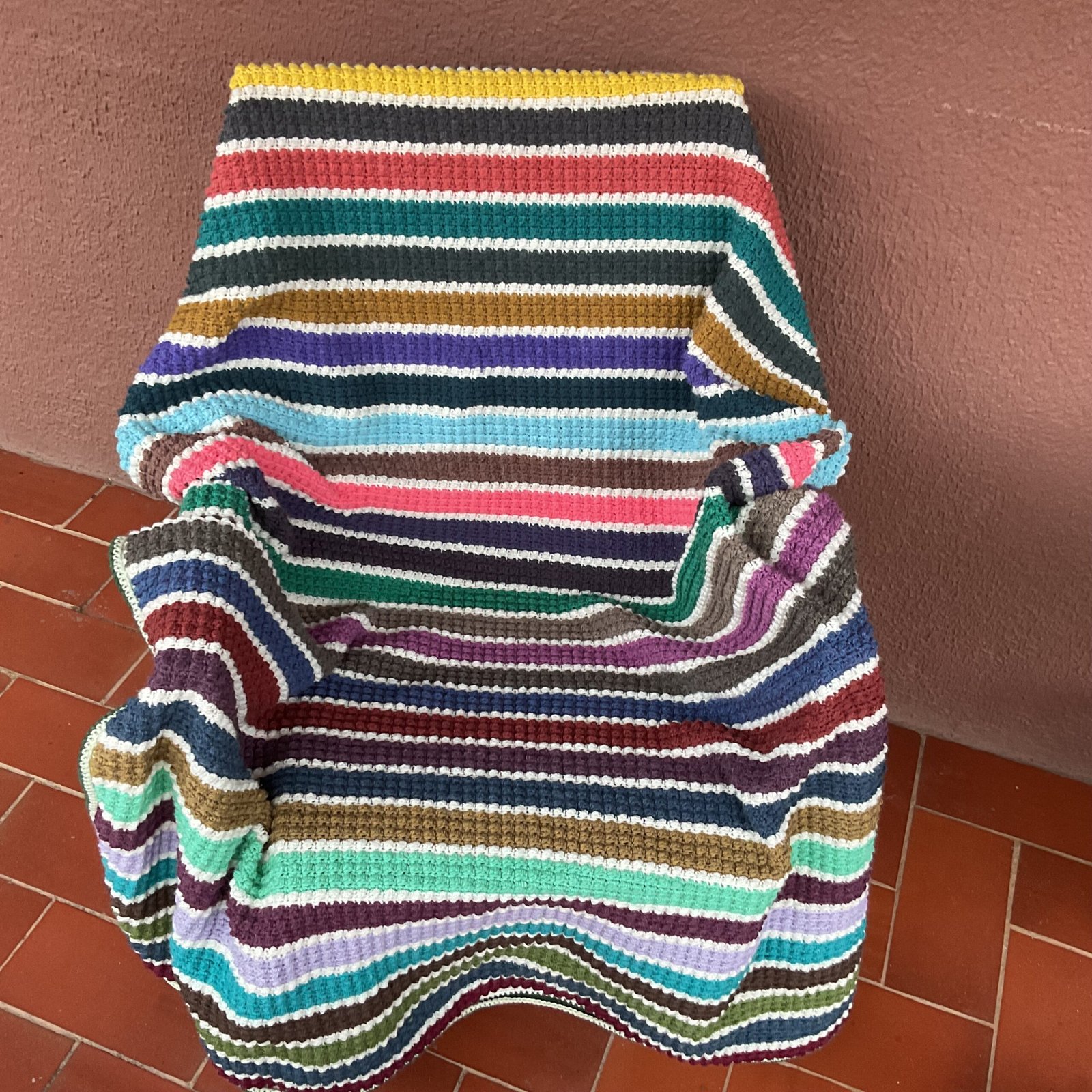 A multi-coloured striped crochet blanket on a chair on a sunny terrace.